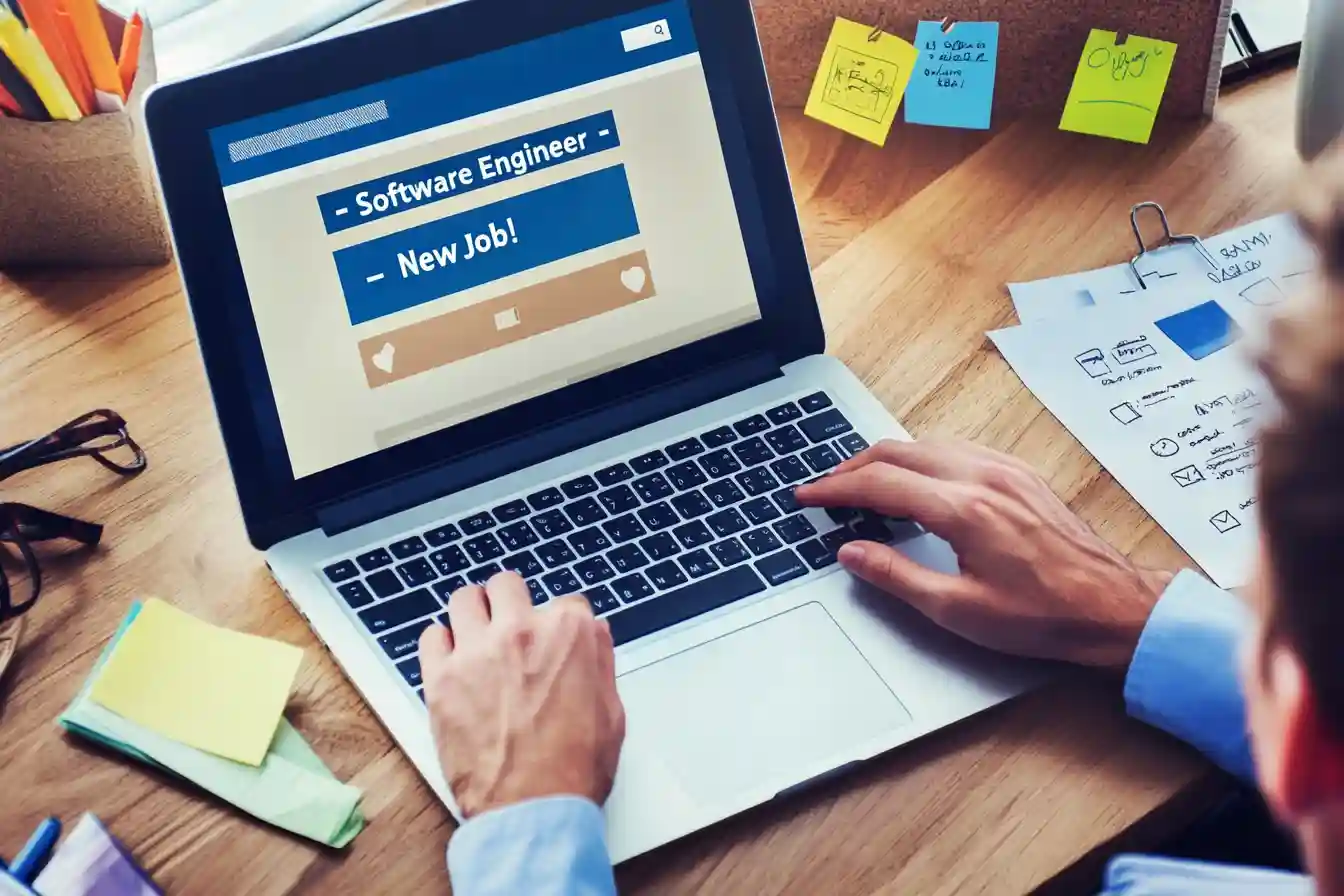 International graduate student adjusts LinkedIn profile on a laptop to reflect a new job, with resume, interview notes, and graduation tassel on the desk.