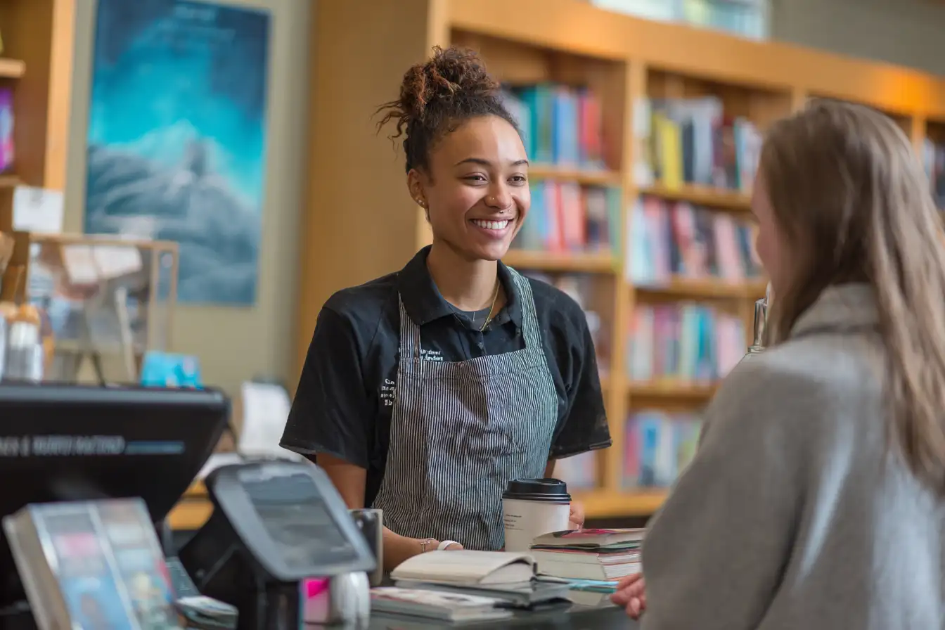 International student working behind the counter at a university café, wearing an apron and serving coffee to a fellow student, with posters for international events in the background.