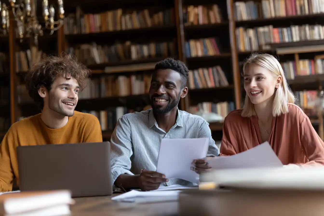Group of international students in a library sharing interview tips and reviewing documents with laptops and notebooks open.