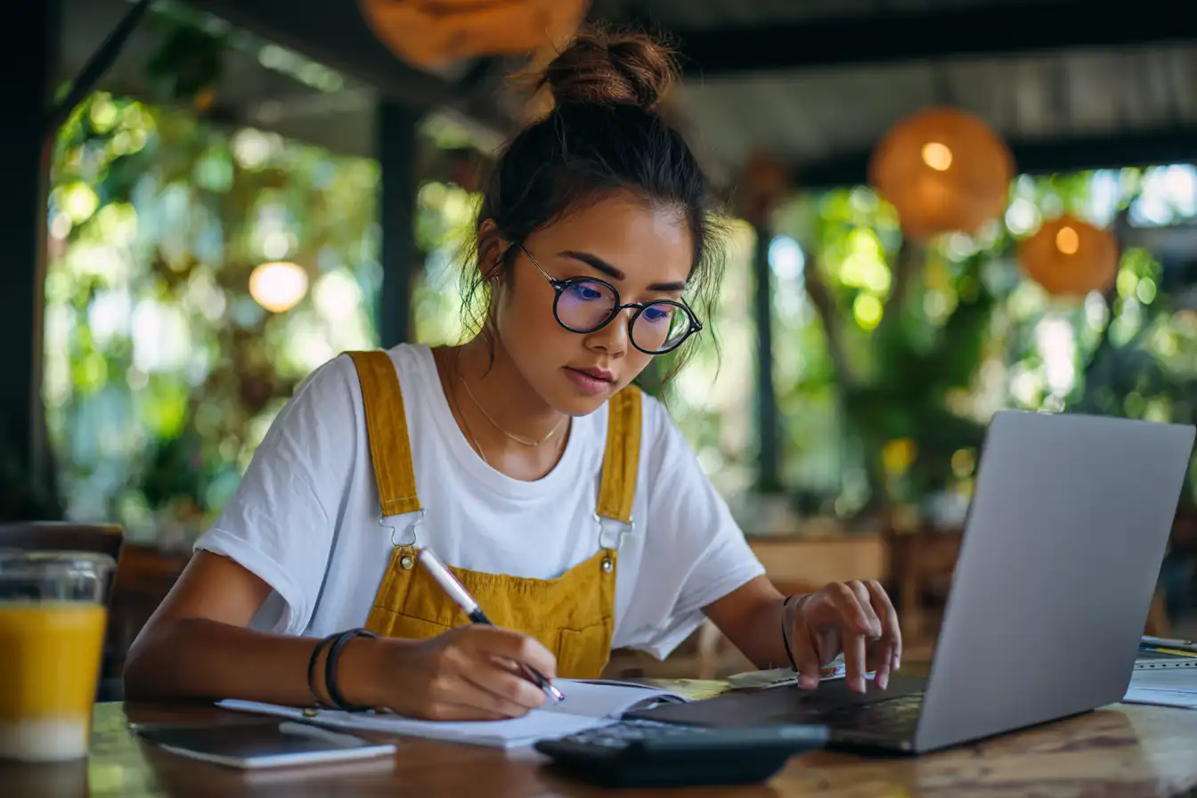 International student in a coffee shop calculating tuition and living costs on a laptop with a maple leaf sticker.