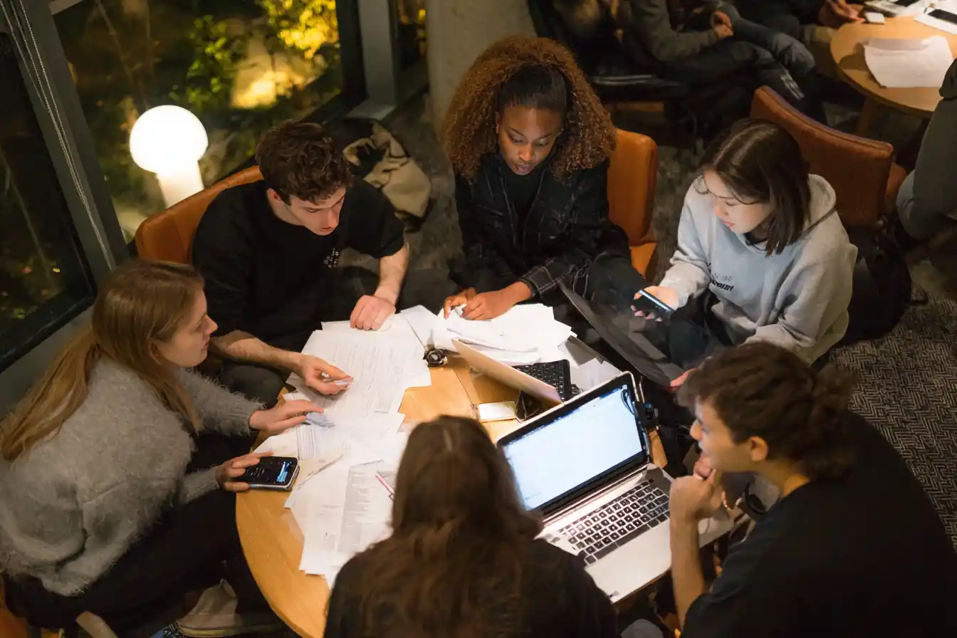 Group of international postgraduates gathered in a common room at night with laptops and paper budgets, discussing monthly expenses and sharing tips on part-time work and visa regulations.
