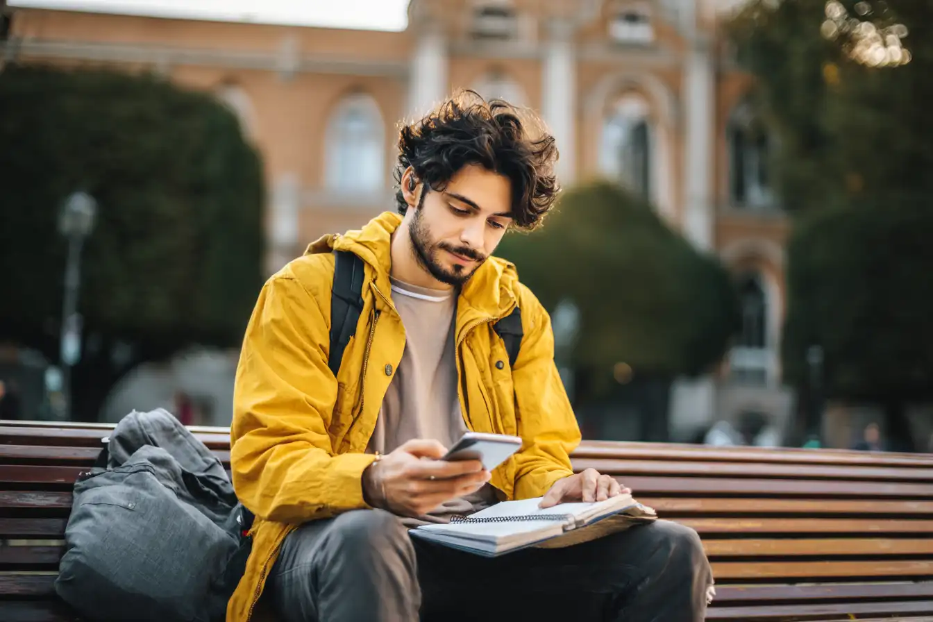 International student seated on a park bench outside a university building, using a smartphone to check loan interest rates while looking thoughtfully at a notebook.