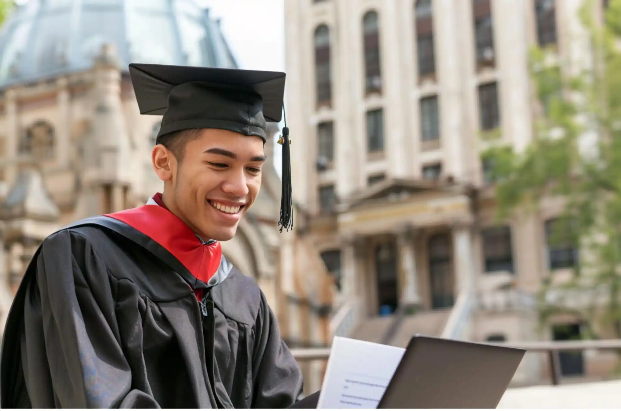 A young international graduate celebrating with a diploma while looking at a laptop showing a job offer.