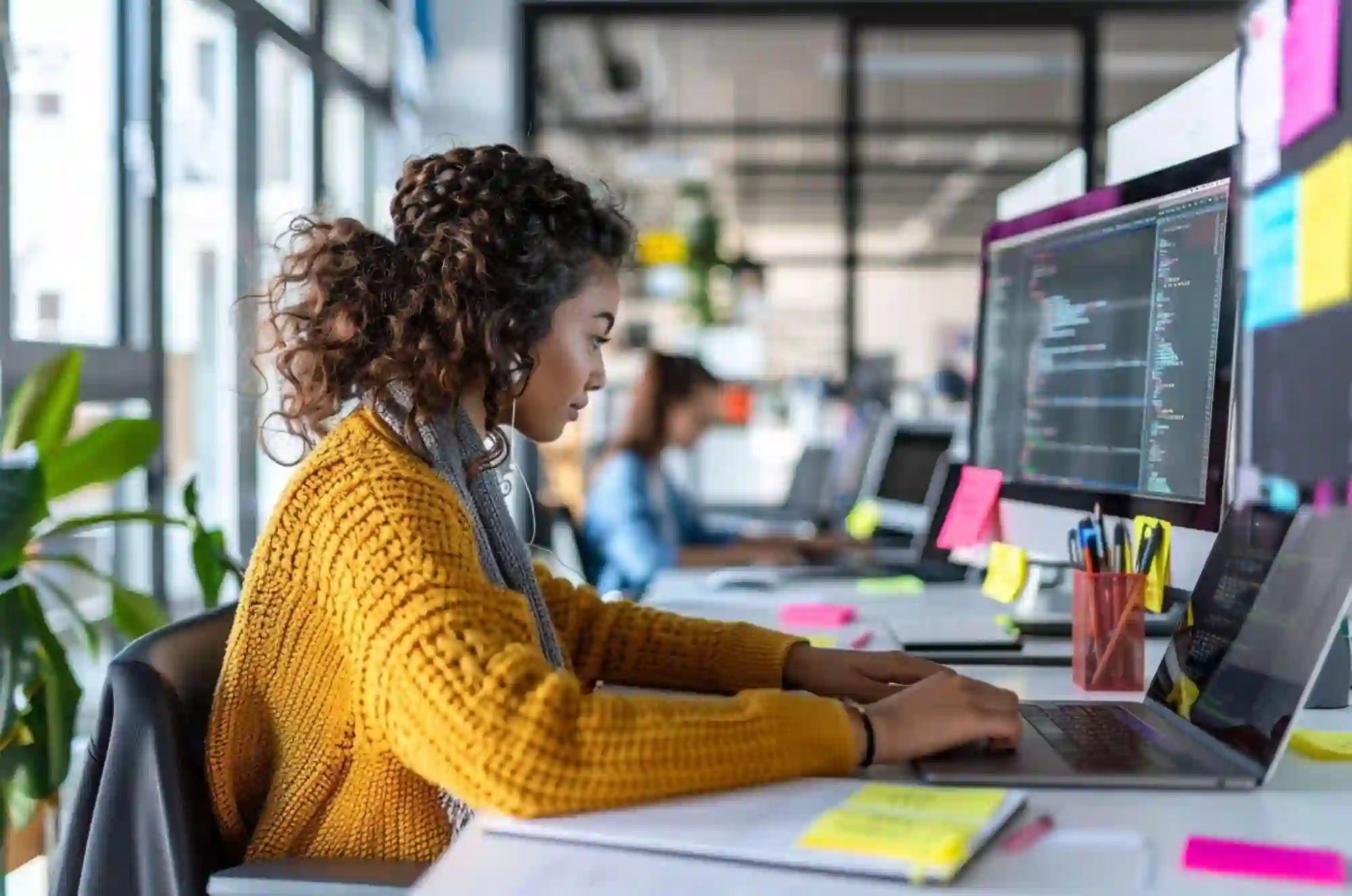 An international student in a modern office with open desks, typing on a laptop while reviewing code on a second monitor. Sticky notes and a planner are on the desk.