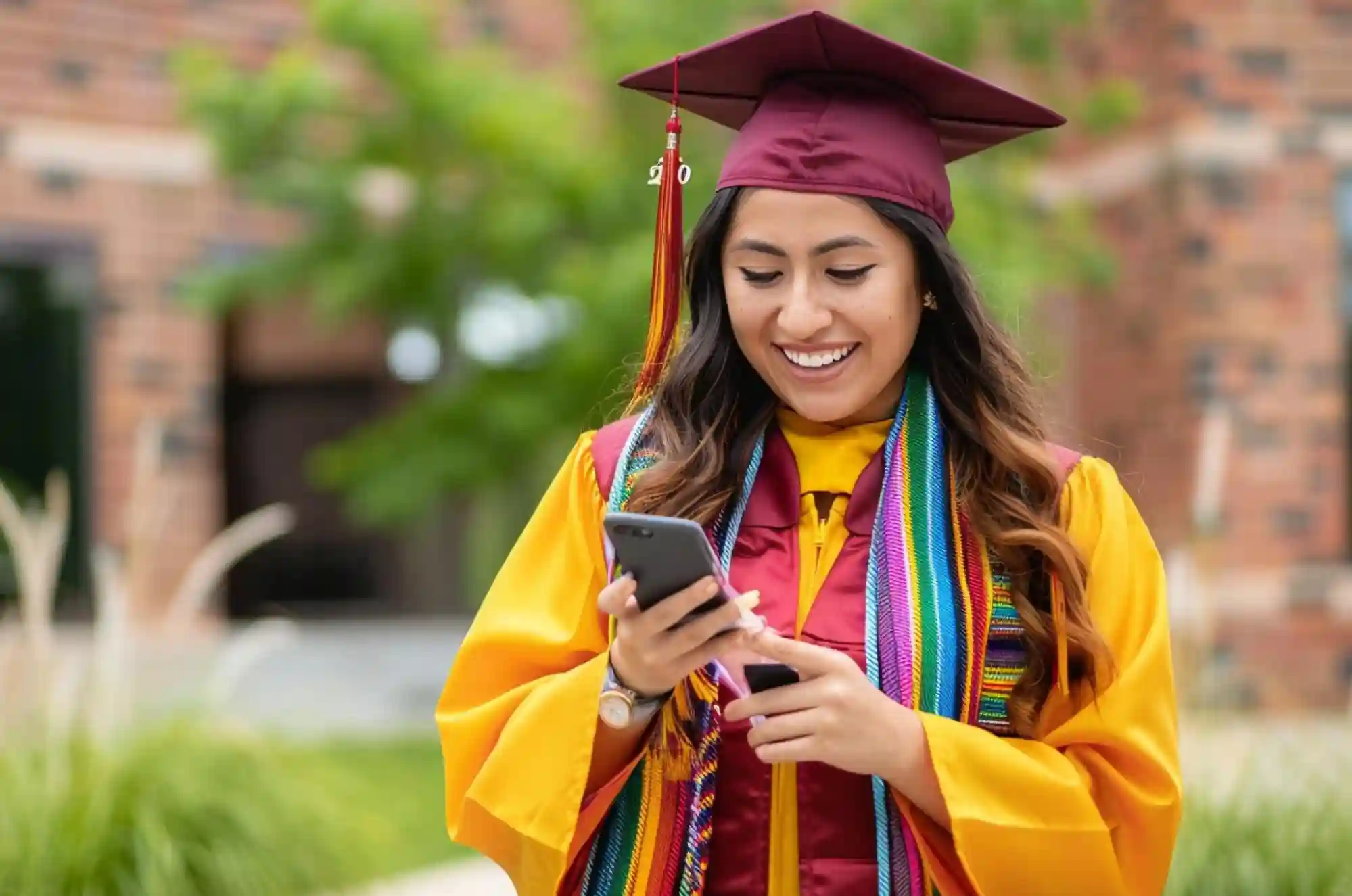 A smiling Columbian student wearing a graduation gown and cap while looking at her phone