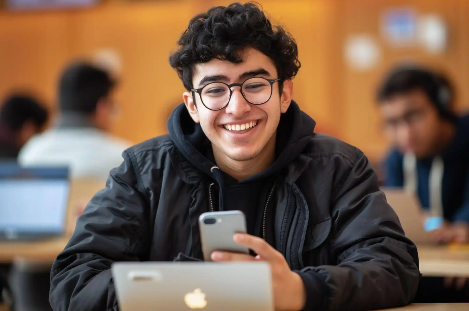 A student smiles warmly while holding a phone in front of a laptop.