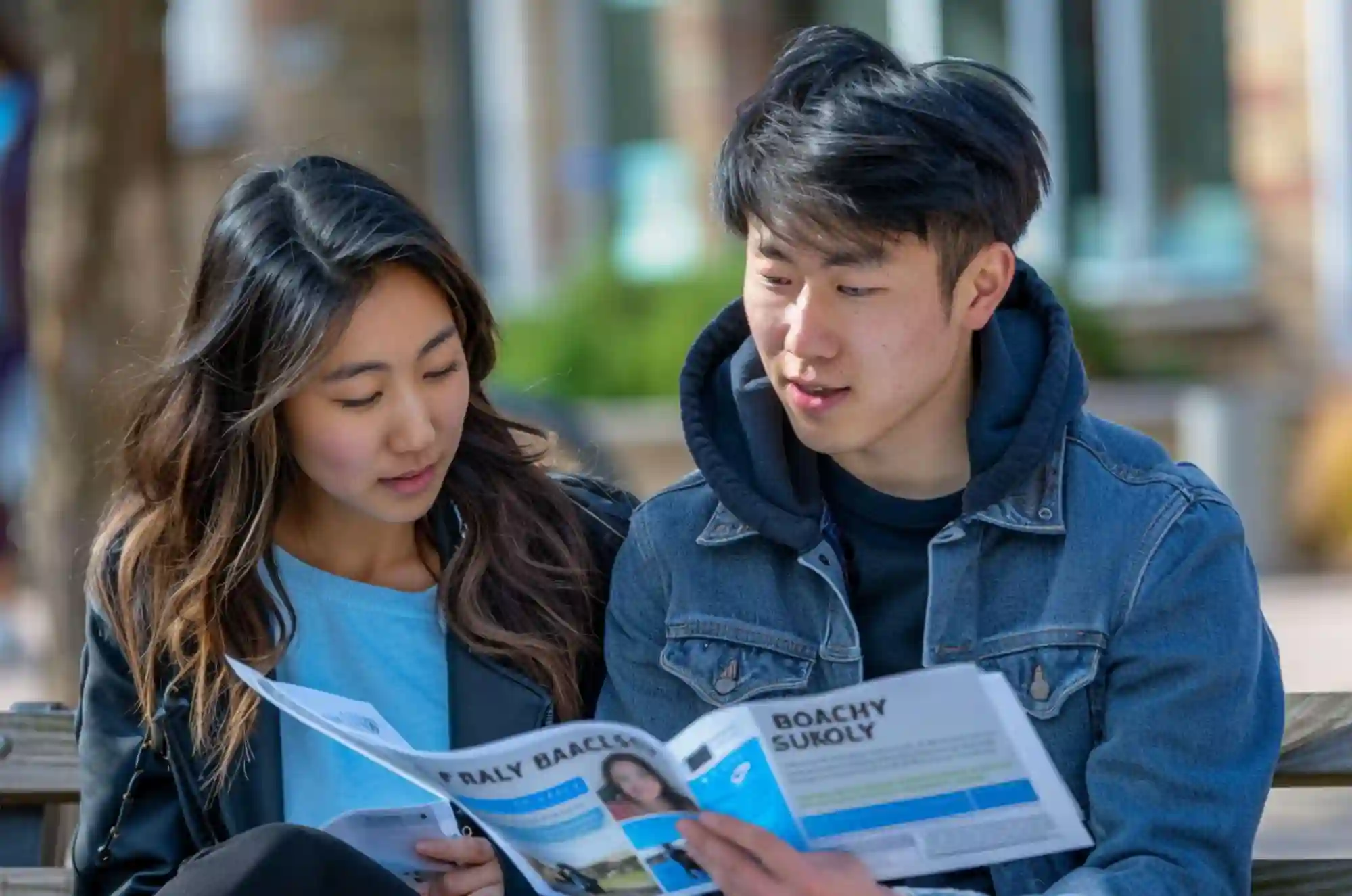 Two international students review a printed bank brochure while seated on a campus bench