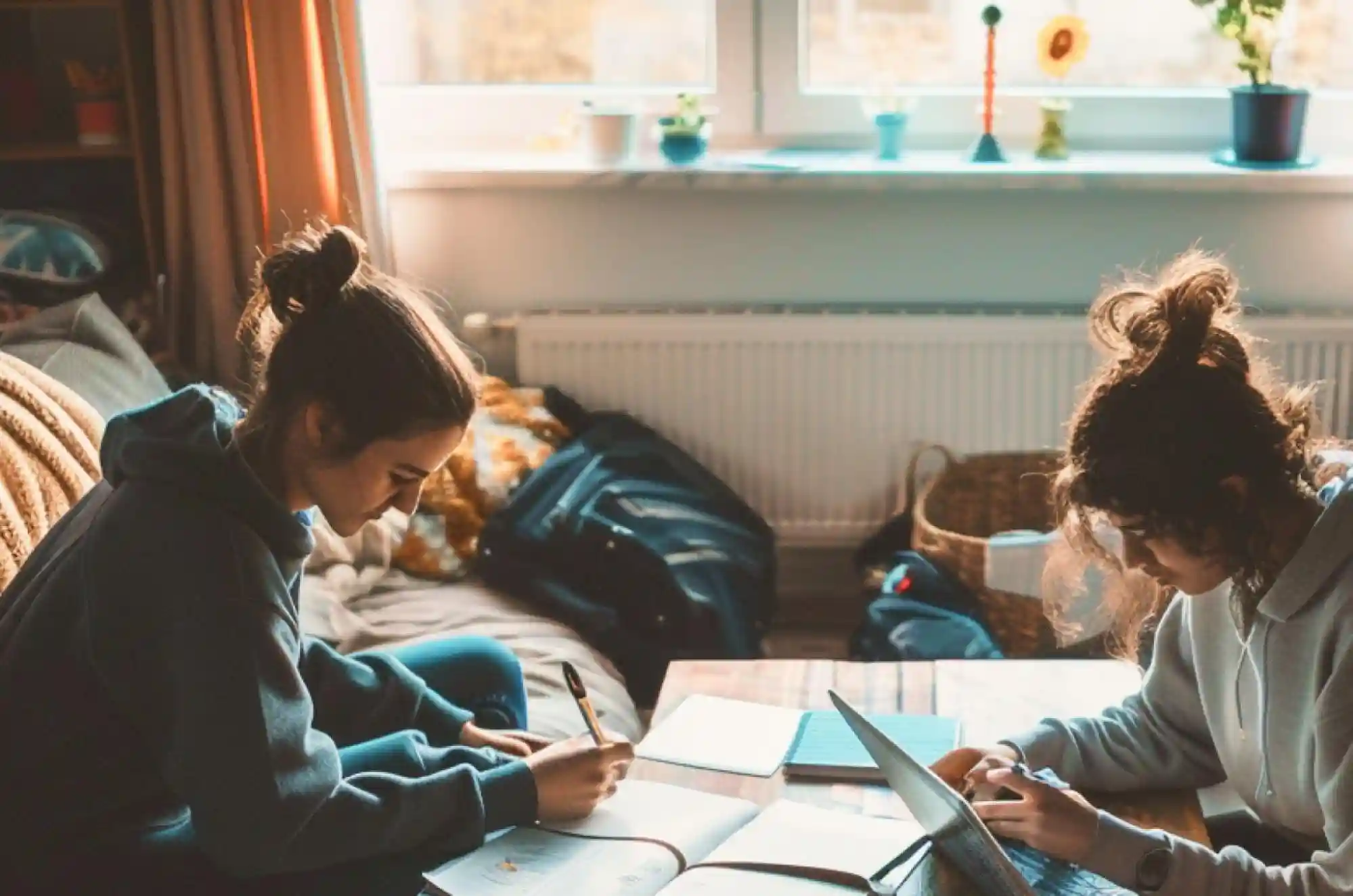 Two international students sit together in a dorm room, one guiding the other through an online bank setup on a laptop.Around them is a lease agreement and an open backpack with folders and receipts.