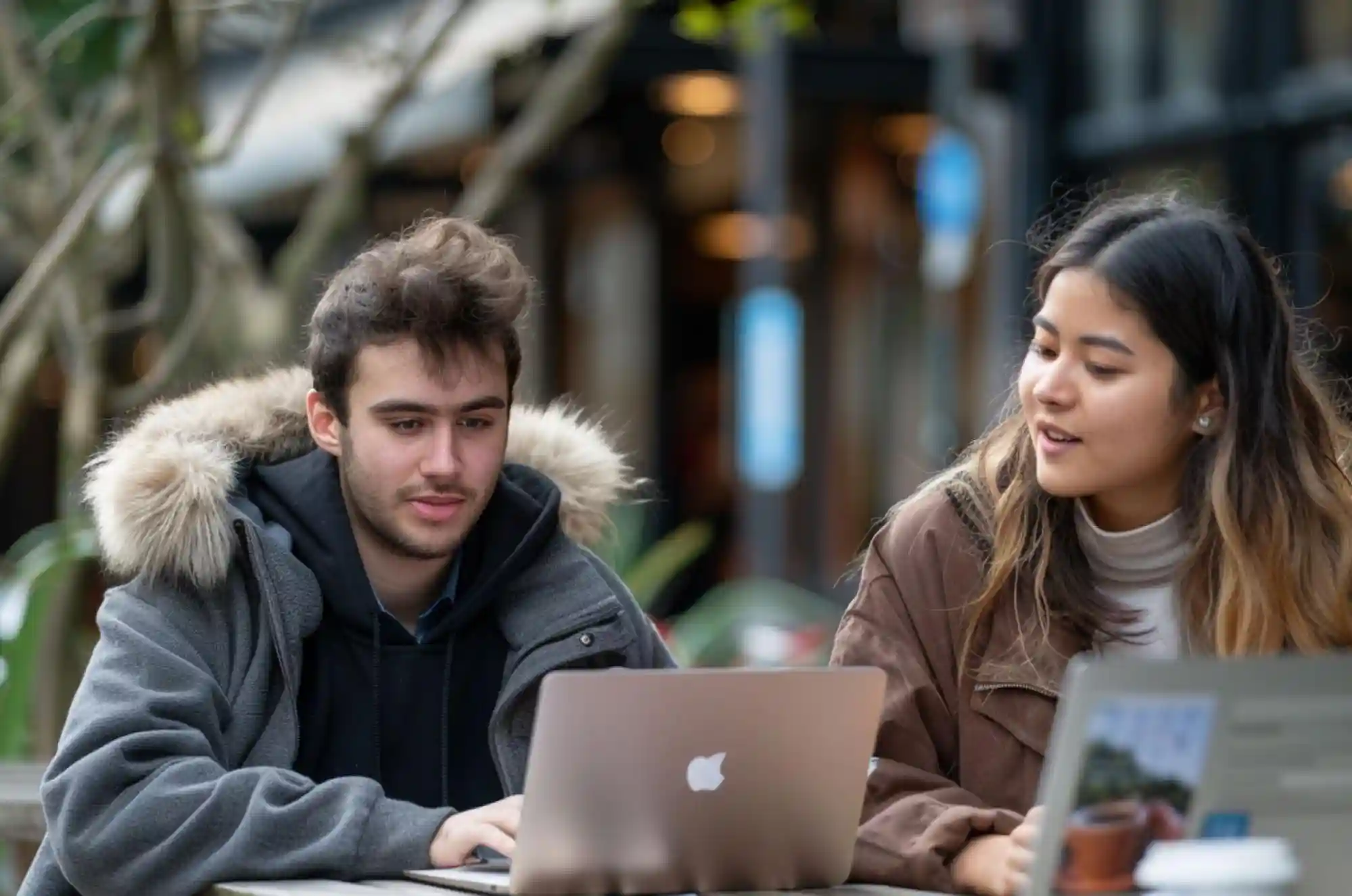 Two students chat over coffee at an outdoor café while reviewing a credit card bill. Backpacks and laptops sit nearby, reinforcing the setting as a break between classes or study sessions.