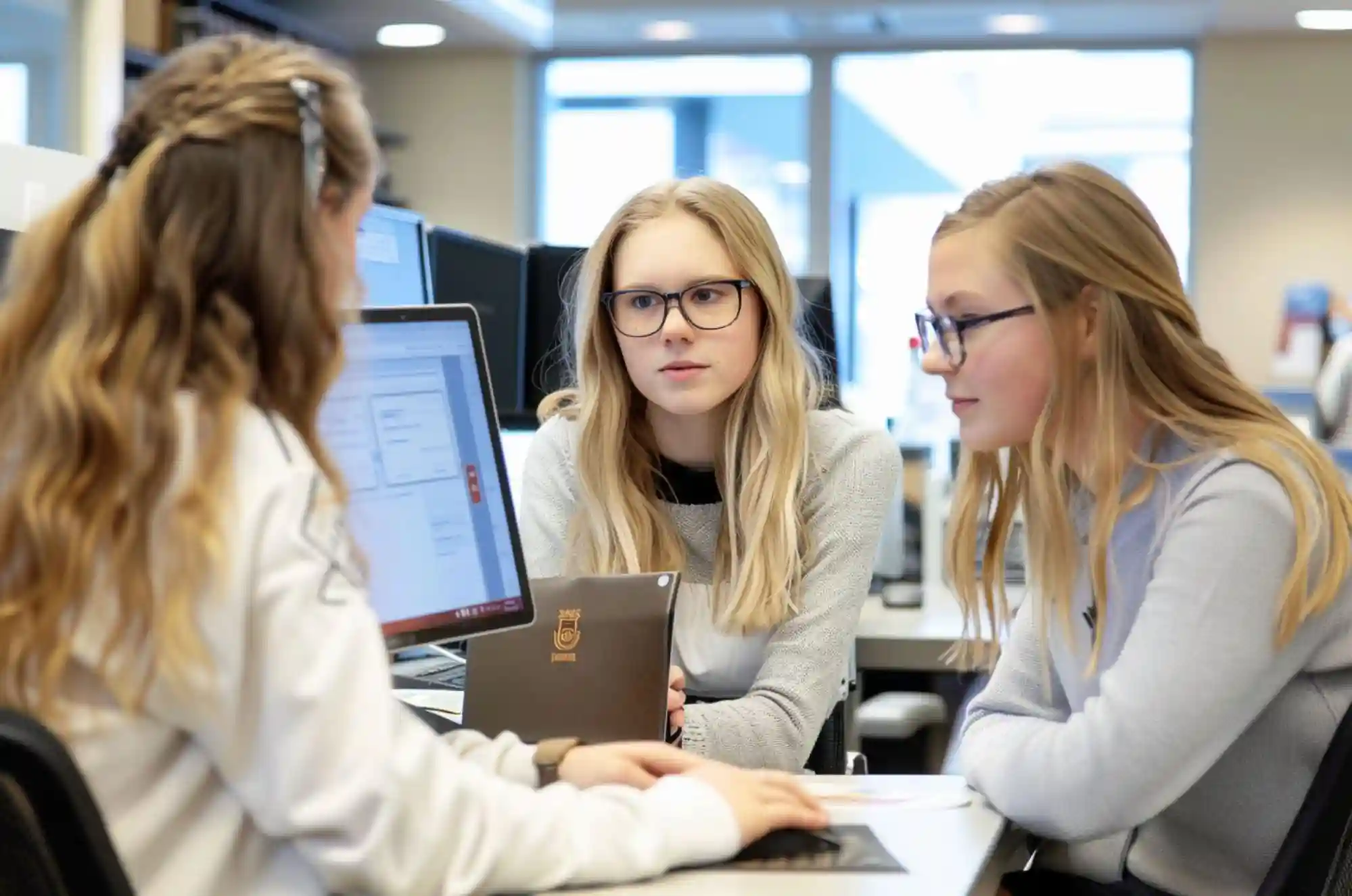 Two students sit at a bank help desk with an advisor.A small table between them includes application forms.