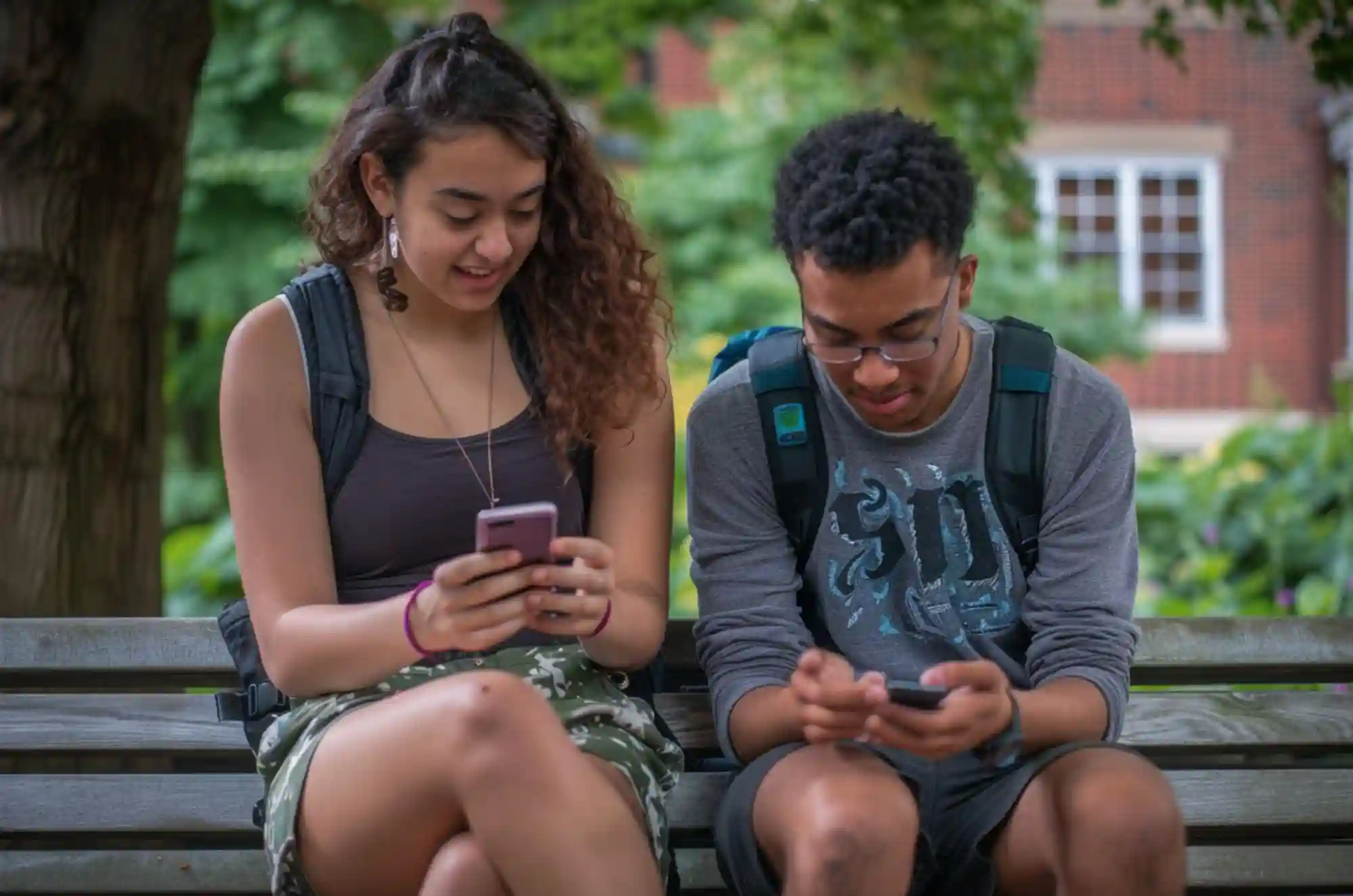 Two students sit on a bench on a university campus comparing mobile apps.