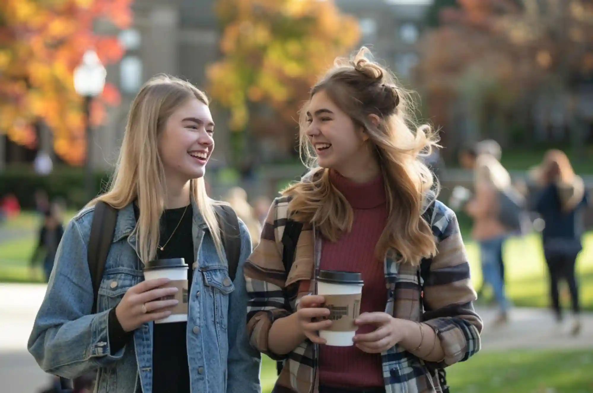 Two students walk across a busy campus quad between classes, holding coffee cups and chatting. The setting captures the everyday rhythm of university life in the U.S.